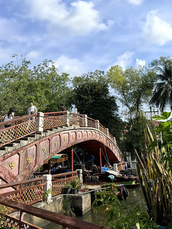 在Khlong Damnoen Saduak运河附近游览时，一艘带顶篷的木船在一座华丽的桥下运送游客，游船行驶在通往Tha Chin和Mae Klong河的水路上